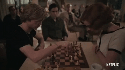 Imagem em movimento em que um homem branco, de cabelos loiros e camiseta preta, movimenta peças de xadrez ao jogar contra uma mulher branca, de cabelos curtos e avermelhados, vestida com blusa branca e preta.