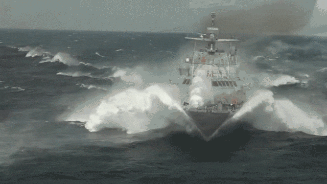 Imagem em movimento em que uma embarcação de porte grande, de cor cinza e aspecto militar, navega em velocidade no mar, produzindo ondas e espuma branca.
